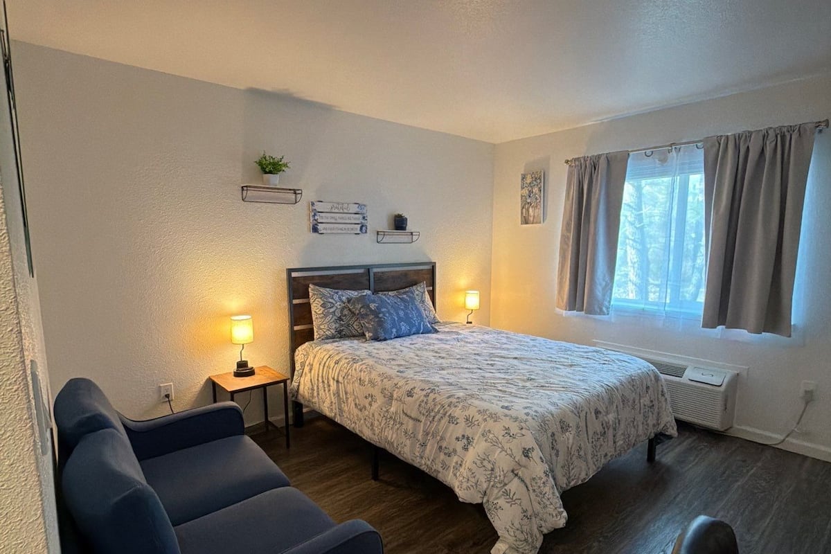 Een slaapkamer met een bed, een bureau, een stoel, een sofa, een nachtkastje met een lamp en een raam met gordijnen.