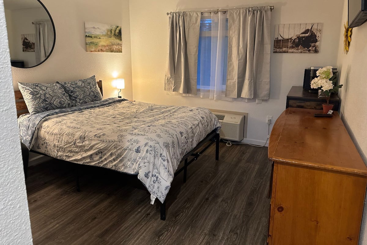 Een slaapkamer met een bed, een nachtkastje, een spiegel, een raam met gordijnen en een kleine plant op de kaptafel.