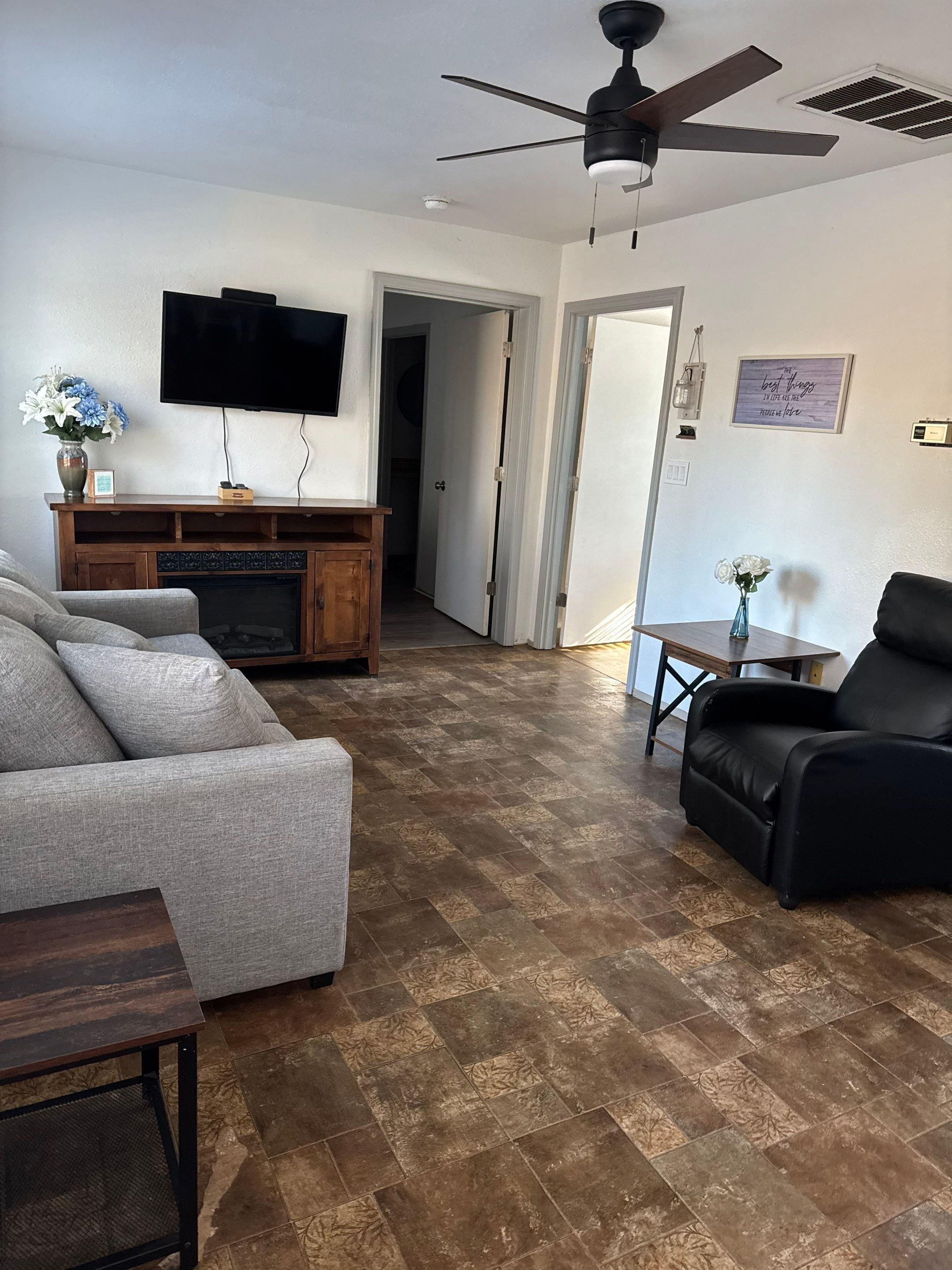 Una sala de estar con un sofá gris, un sillón de cuero negro, un mueble de madera para entretenimiento y un televisor de pantalla plana montado en la pared.