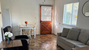 A living room with a gray sofa, a dining table with chairs, a black leather chair, a wooden coffee table, and a vase with white flowers. There is a wooden door with a window and blinds, a ceiling fan, and a window with blinds. - Brownsville Motel (Brownsville)