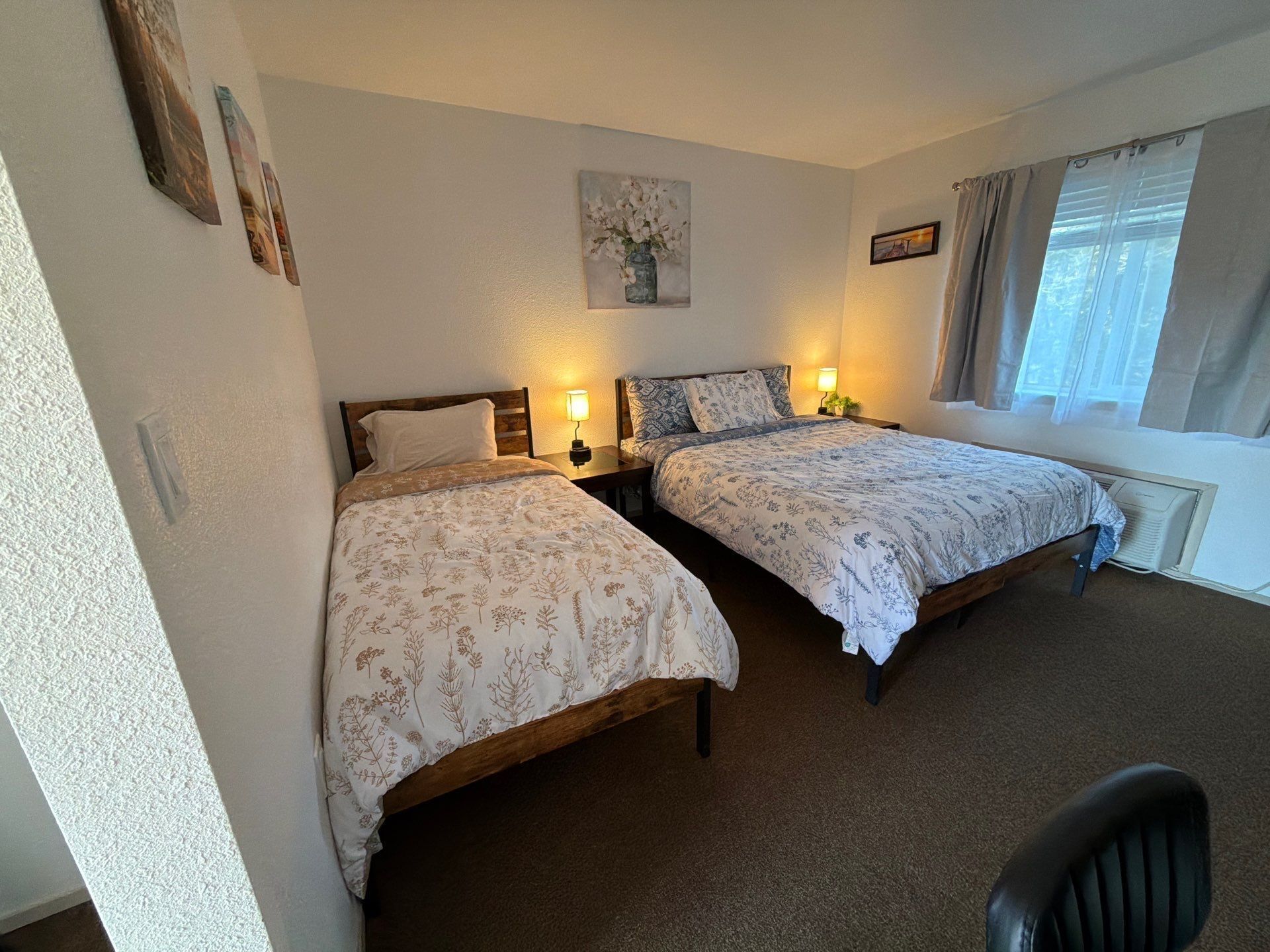 Quarto com duas camas, cada uma com uma mesa de cabeceira e uma luminária. Há uma janela com cortinas, um quadro na parede e um radiador sob a janela.