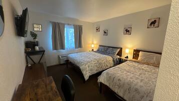 Ein Zimmer mit zwei Betten, einem Schreibtisch, einem Stuhl, einem an der Wand befestigten Fernseher und einem Fenster mit Vorhängen.