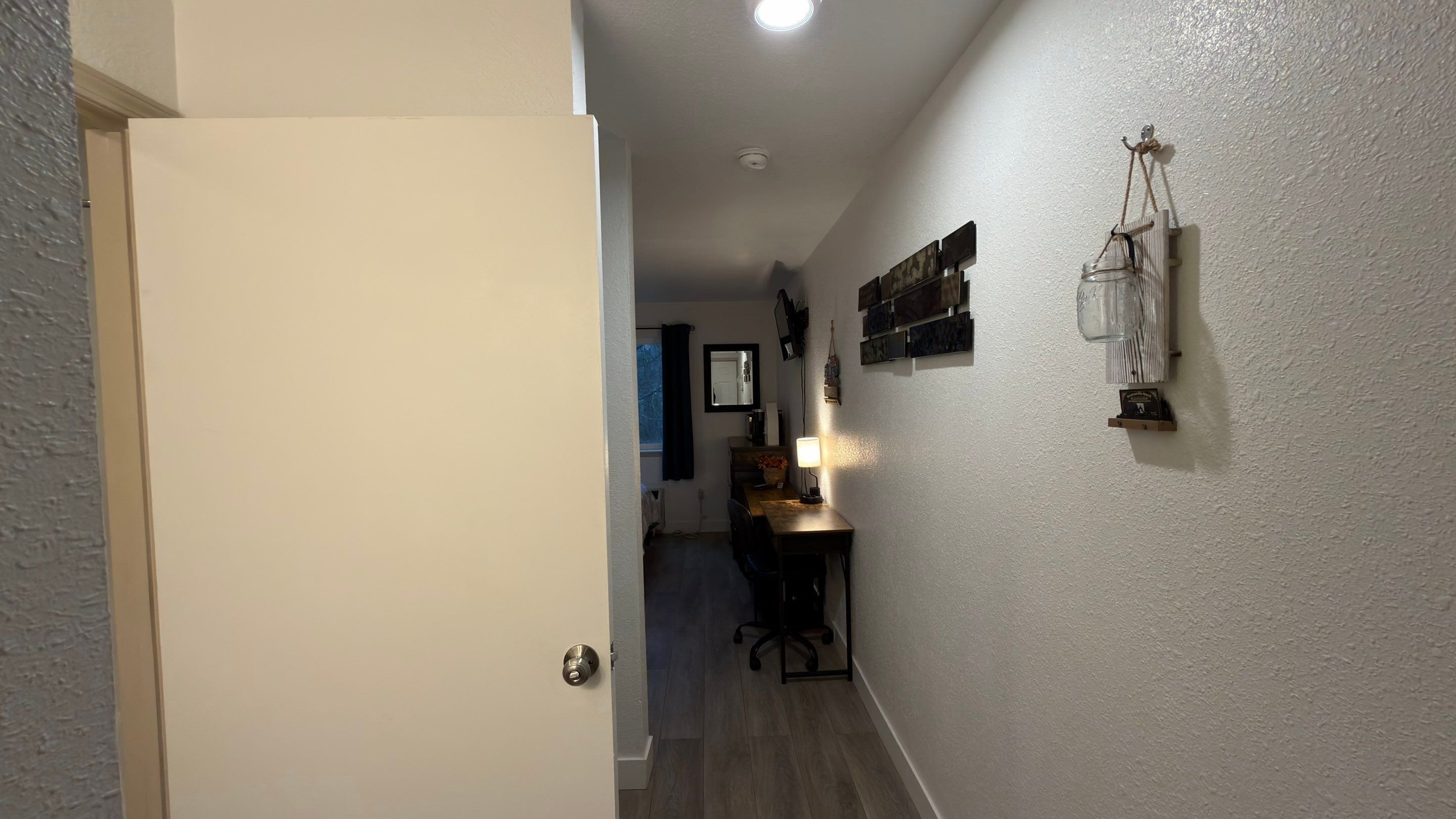 Un couloir avec une porte en bois, un bureau avec une lampe et un miroir accroché au mur.