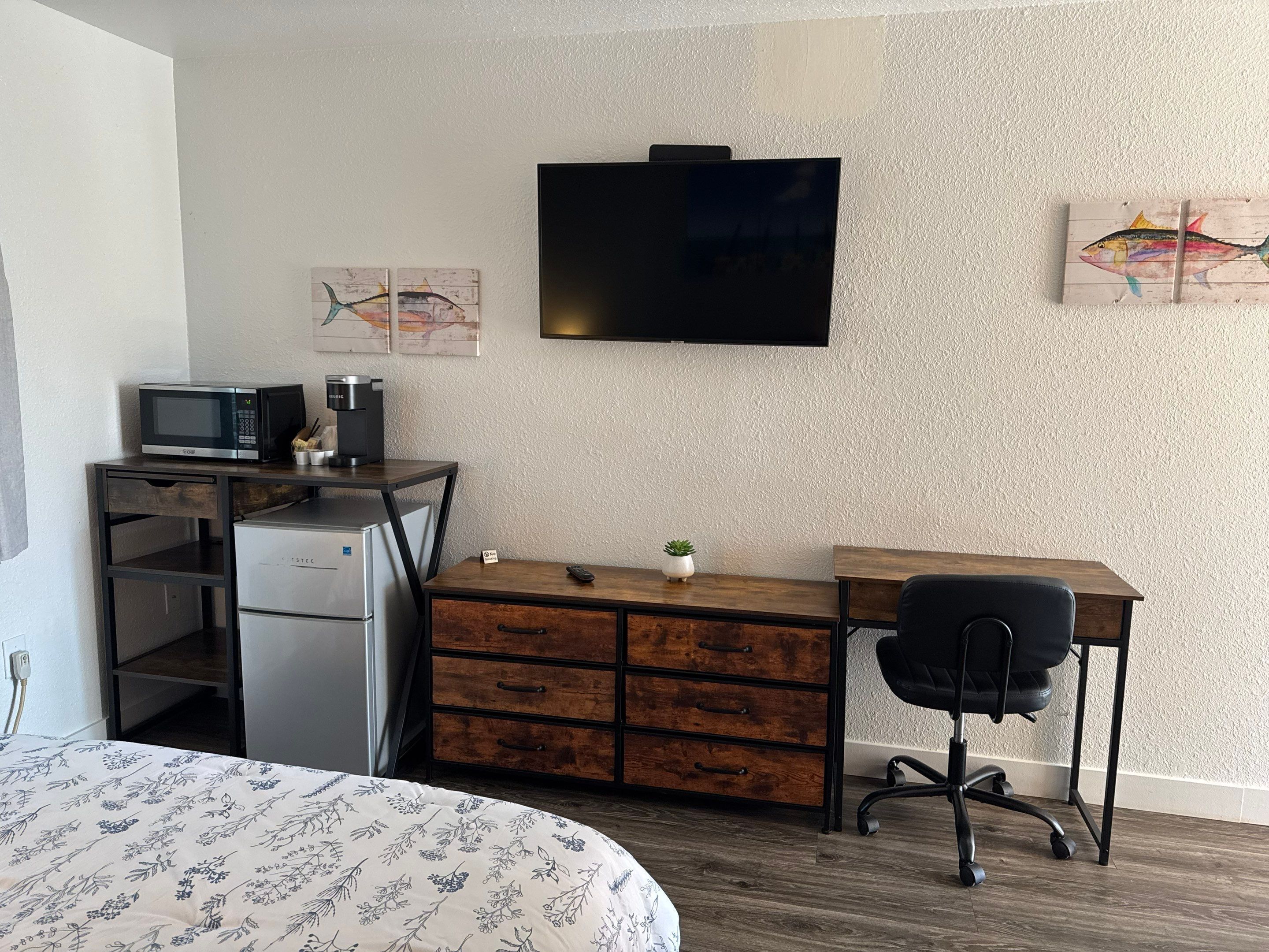 Ein kleines Hotelzimmer mit einem Bett, einem Schreibtisch mit Computer, einer kleinen Kühlschrank, einem Fernseher und Bildern an den Wänden.