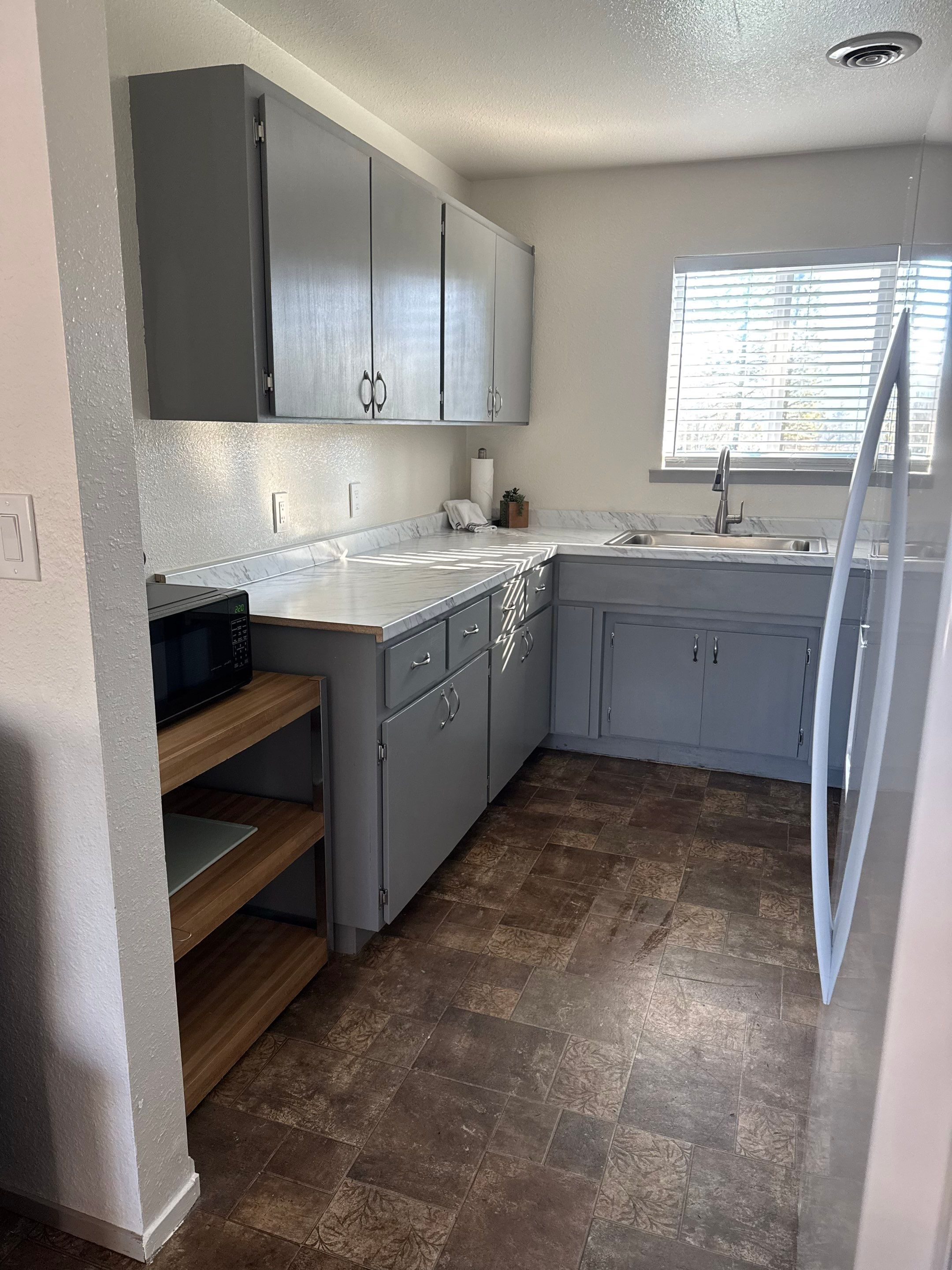 A kitchen with grey cabinets, a microwave, a sink, and a window with blinds.