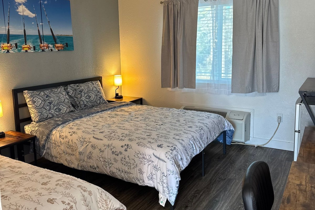 Een slaapkamer met twee bedden, een bureau, een stoel, een lamp, een schilderij en een raam met gordijnen.