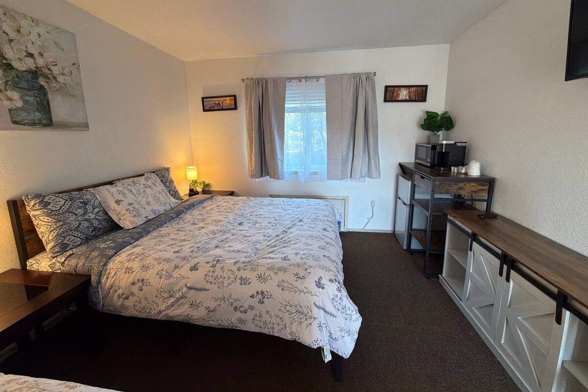 Een slaapkamer met een bed, nachtkastje, lamp, raam met gordijnen en een bureau met een plant.