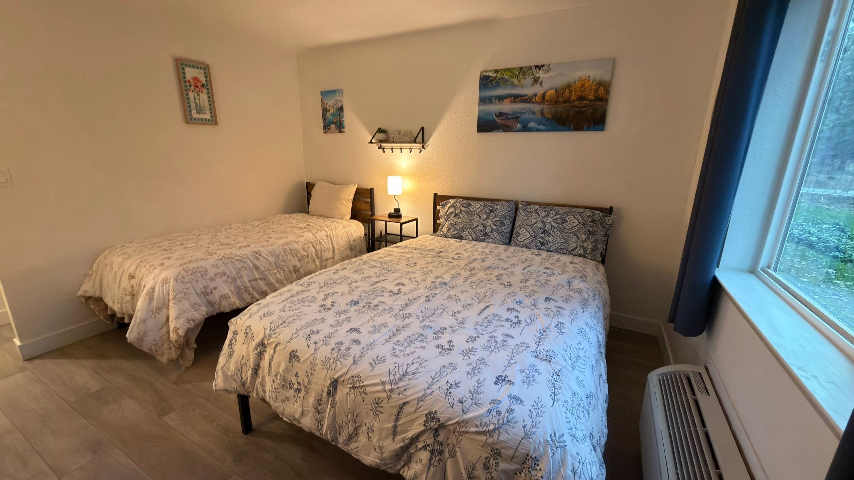 A bedroom with two single beds, a nightstand with a lamp, and a window with a view of greenery.