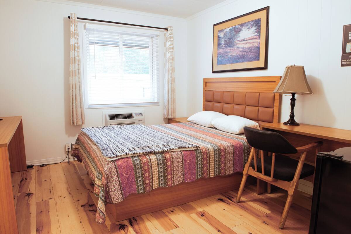 A bedroom with a wooden bed, a desk, a chair, a window with curtains, and a framed picture on the wall.
