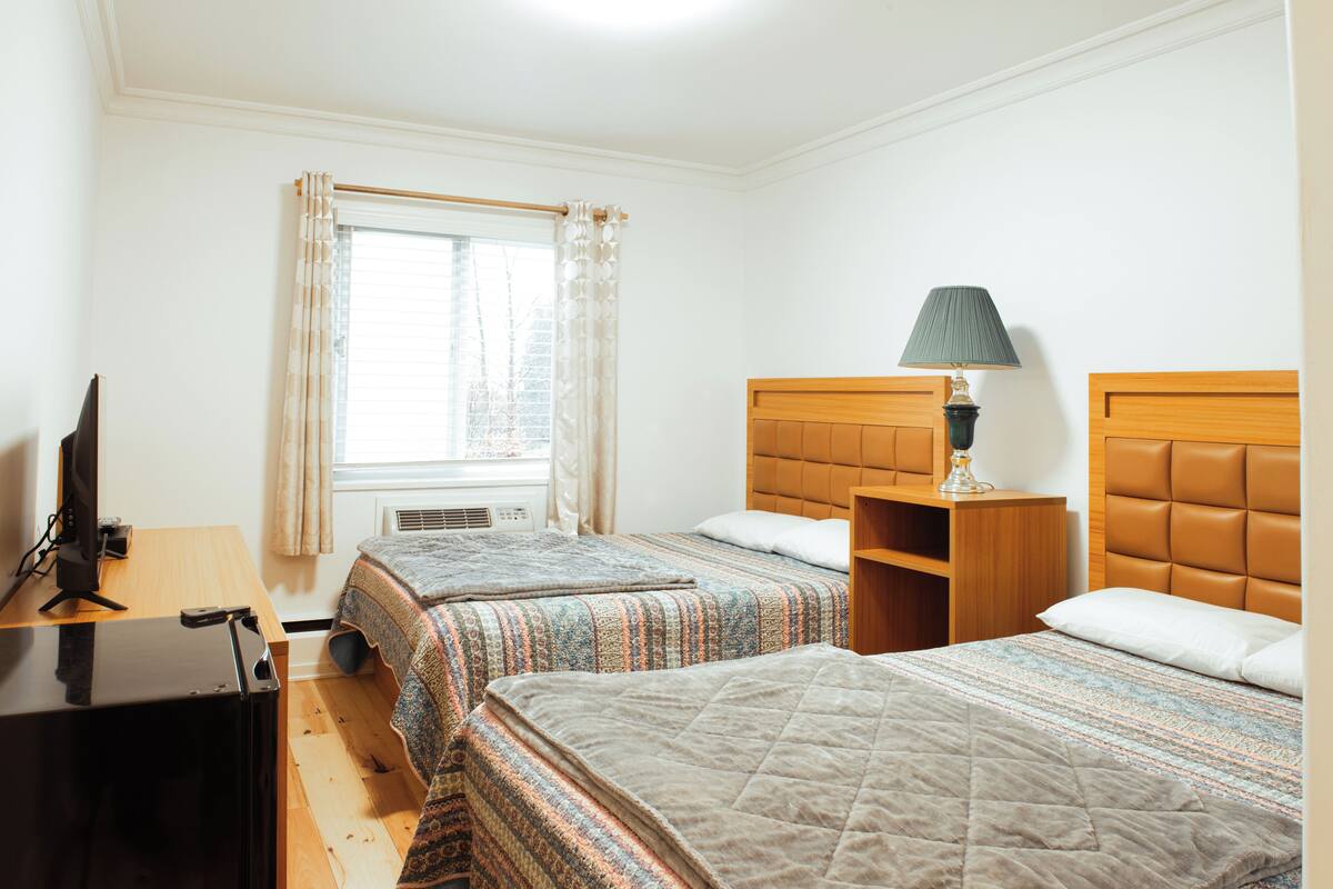A hotel room with two beds, a desk with a computer, a lamp, and a window with curtains.