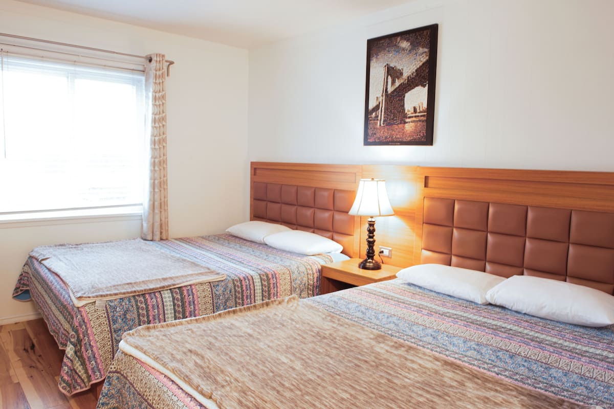 A hotel room with two beds, a wooden headboard, a nightstand with a lamp, a window with curtains, and a framed picture on the wall.