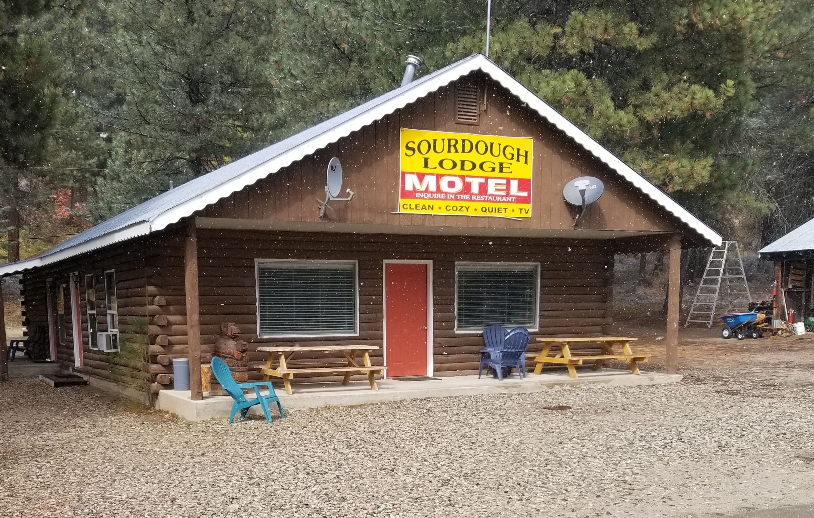 Una baita di legno con un cartello per Sourdough Lodge Motel, caratterizzata da una porta rossa, due finestre e un'antenna parabolica.