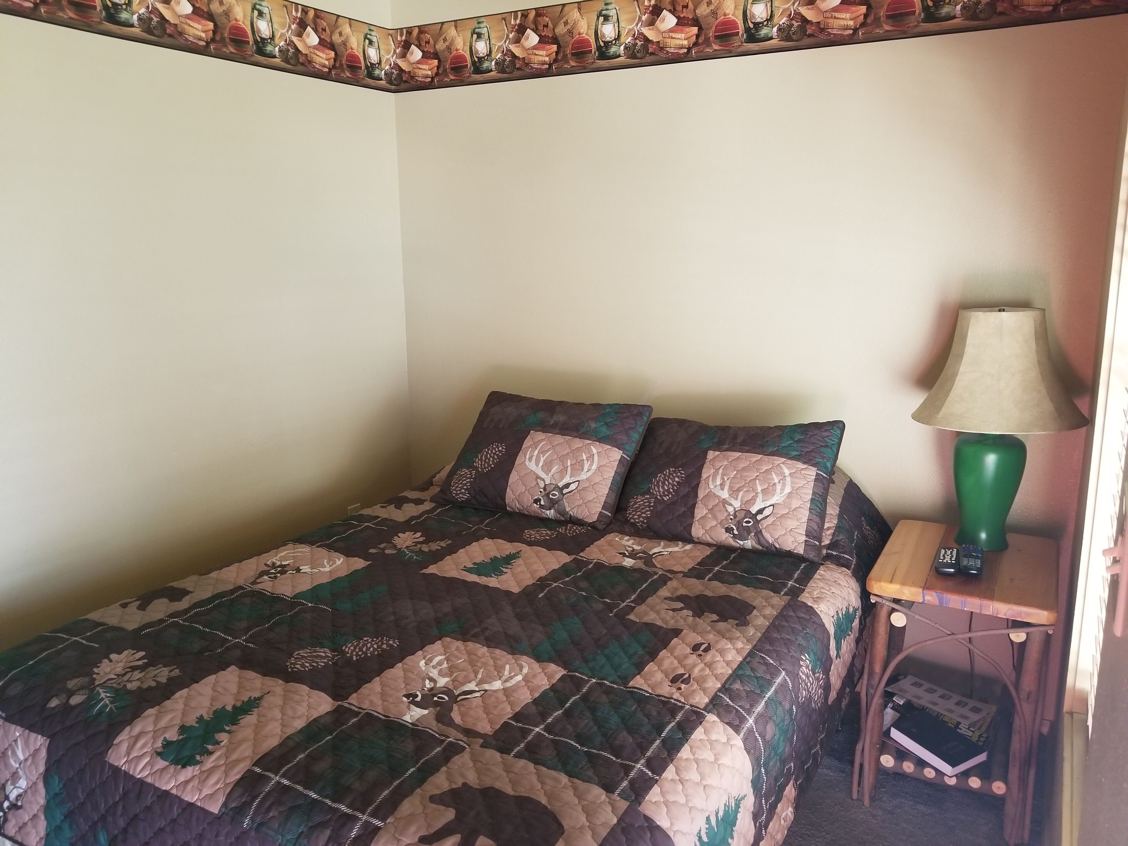 Ein Bett mit einer Quiltdecke, die Hirsche und Kiefern zeigt, eine grüne Lampe auf einem Nachttisch aus Holz und eine Fernbedienung auf dem Nachttisch.