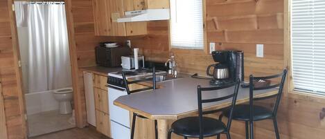 Une cuisine rustique aménagée dans une cabane en bois, avec un îlot central, des meubles en bois, une cuisinière et une fenêtre équipée de stores.