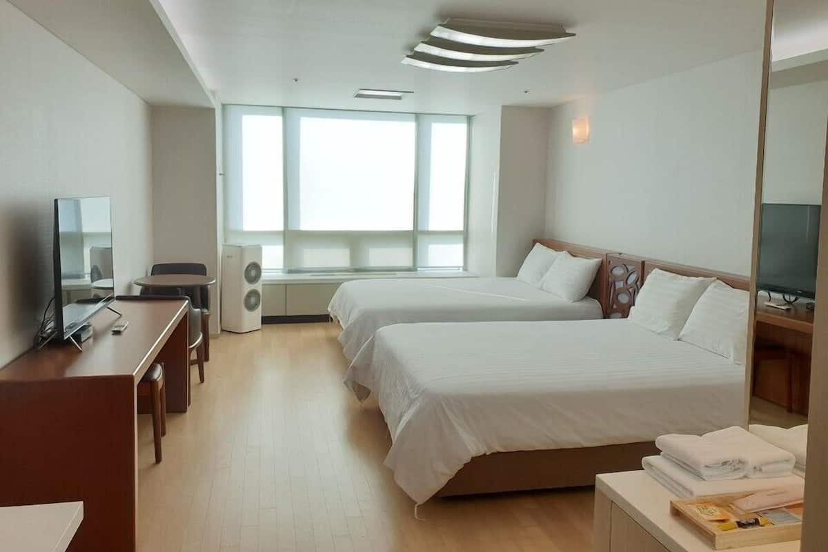 Habitación de hotel con dos camas, un escritorio con silla, un televisor y un ventanal que proporciona luz natural.