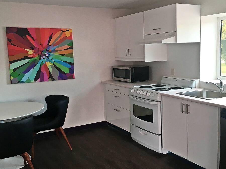 A compact kitchen with white cabinets, a microwave, a stove, and a sink. There's a colorful abstract painting on the wall and a small round table with two chairs.