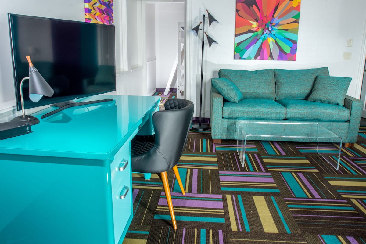A modern living room with a teal desk, a black chair, a teal sofa, and a colorful abstract painting on the wall.