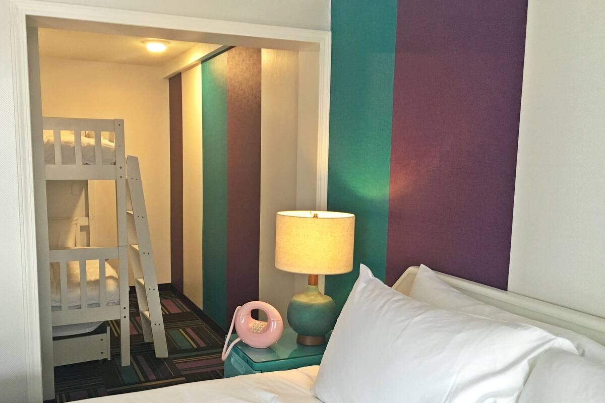 A hotel room with a bed, a nightstand with a lamp, a telephone, and a ladder leading to a bunk bed.