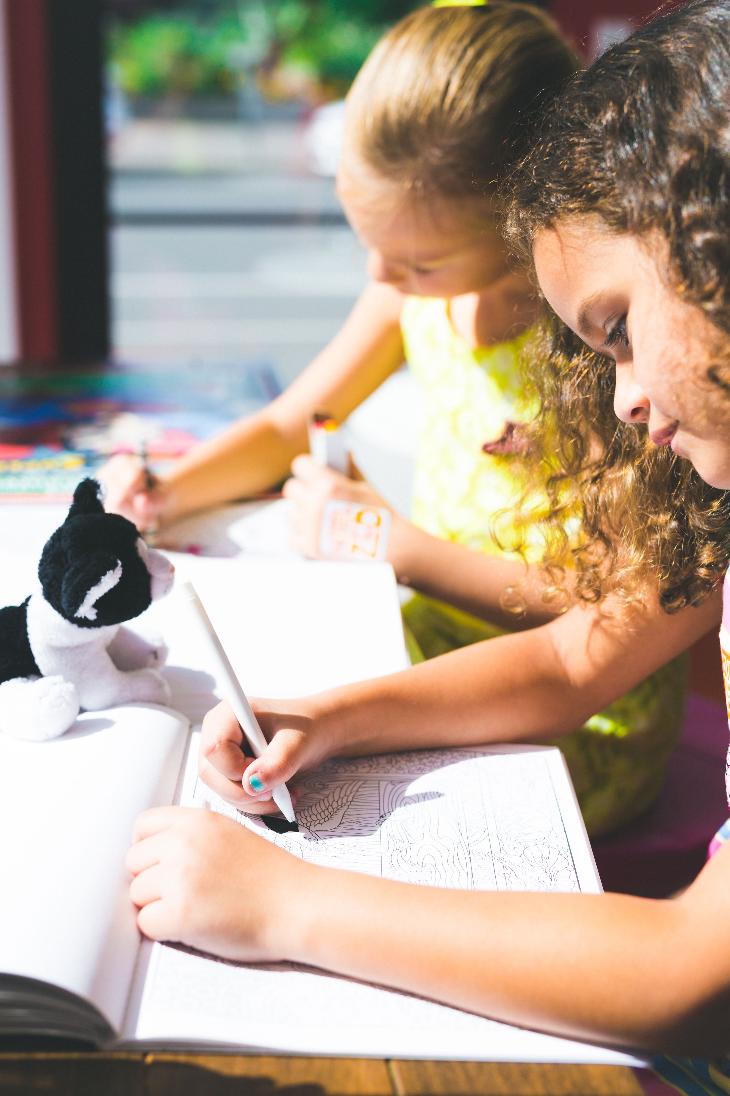 Dos niños dibujando en una mesa con un libro y un peluche.