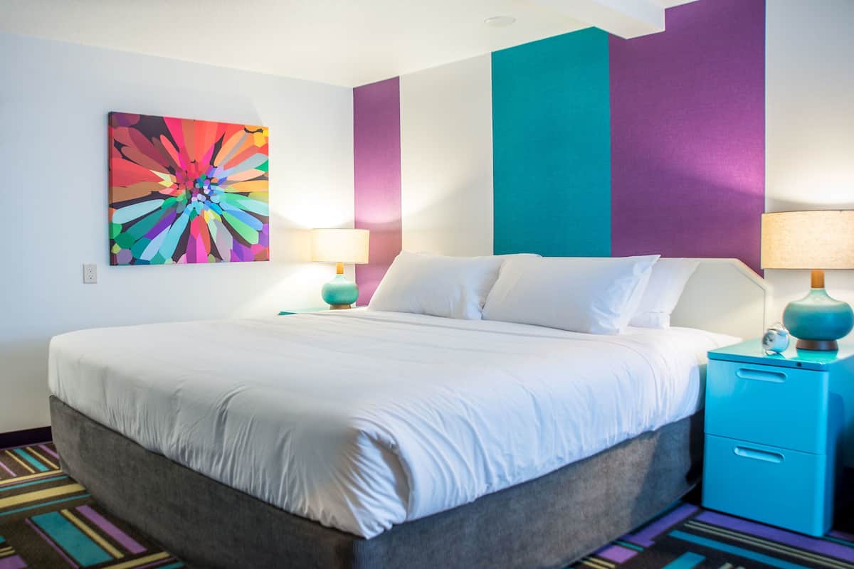 A modern hotel room with a large bed, two bedside tables with lamps, a colorful abstract painting on the wall, and a striped carpet.