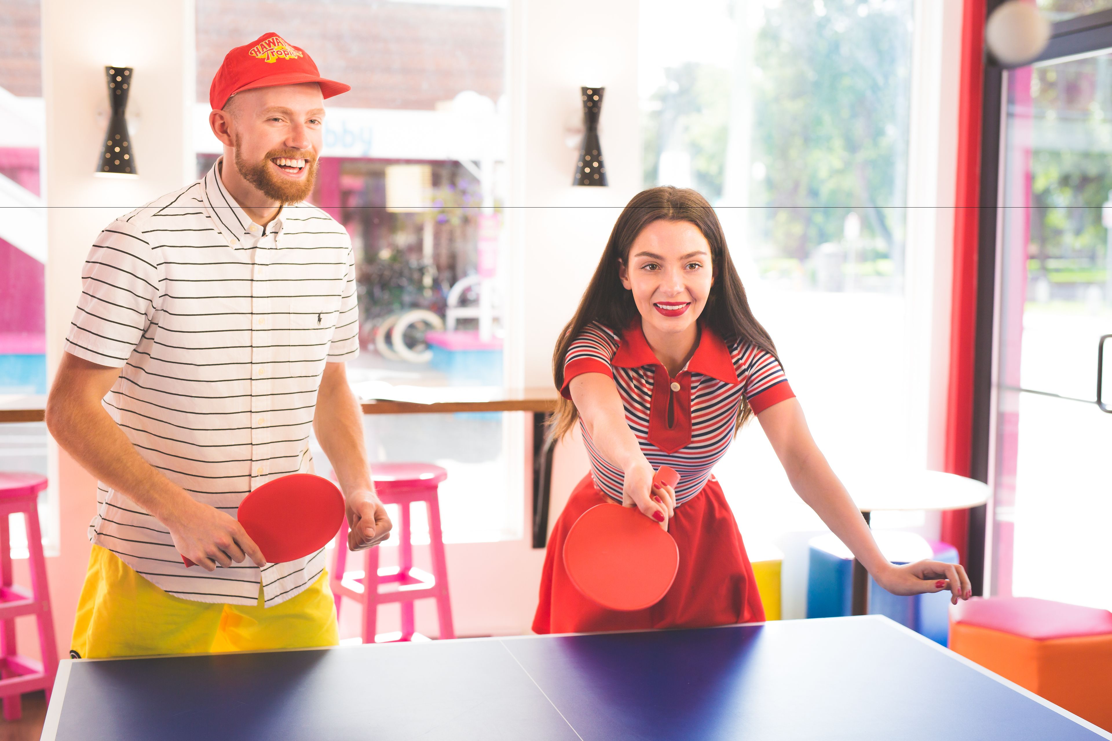 Two people playing table tennis in a brightly lit room with colorful stools and a table.