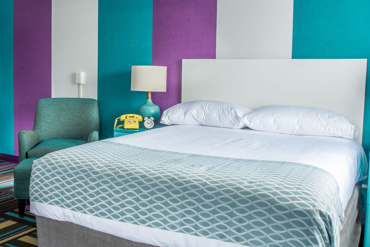 A hotel room with a bed, a chair, a nightstand with a lamp, and a telephone.