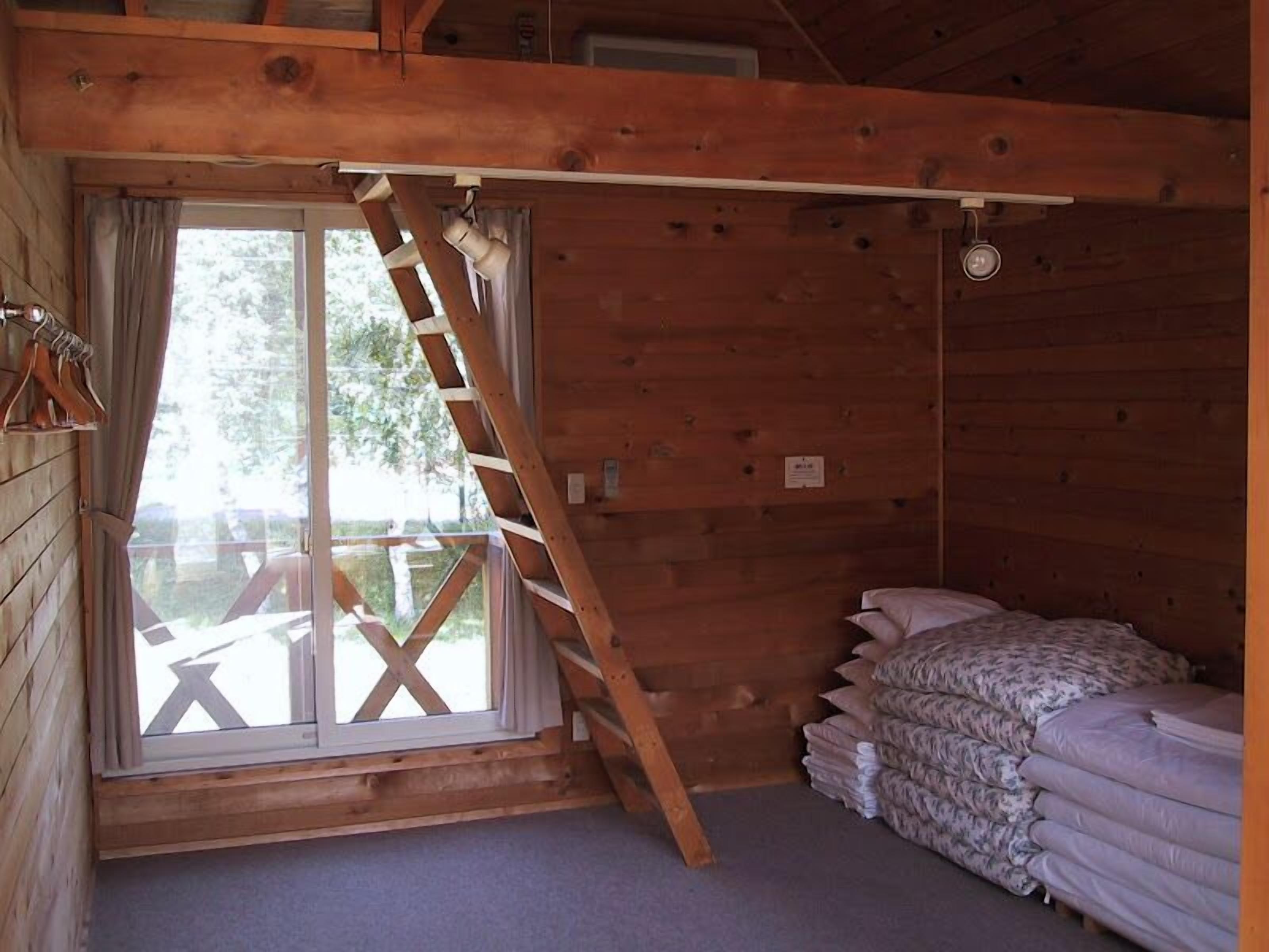 Una habitación tipo cabaña de madera con cama en altillo, escalera y ventana con vista a una terraza.