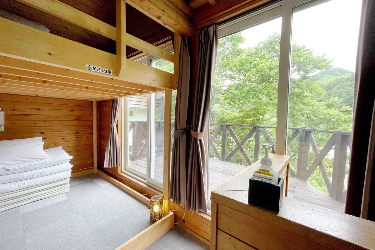 A wooden cabin room with a bed, a small table with a bottle, and a window with a view of greenery.
