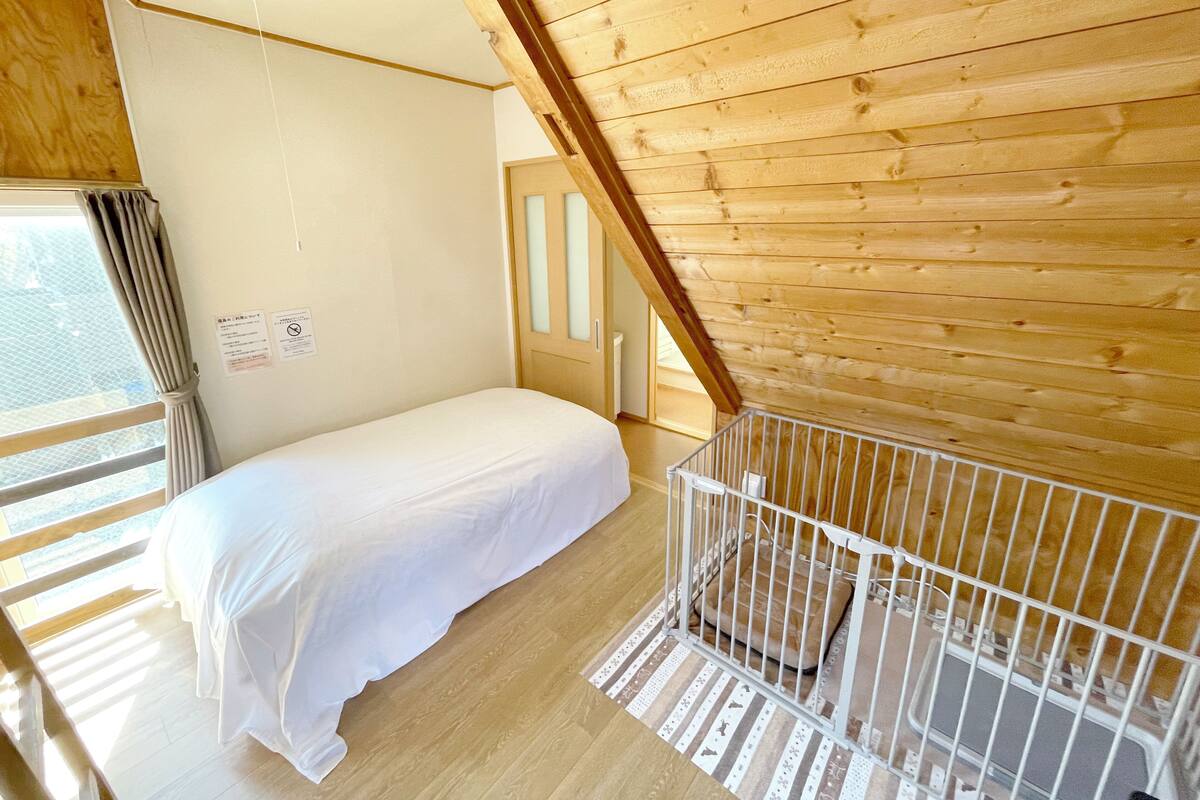 A room with a bed, a crib, wooden ceiling beams, a window with curtains, and a door.