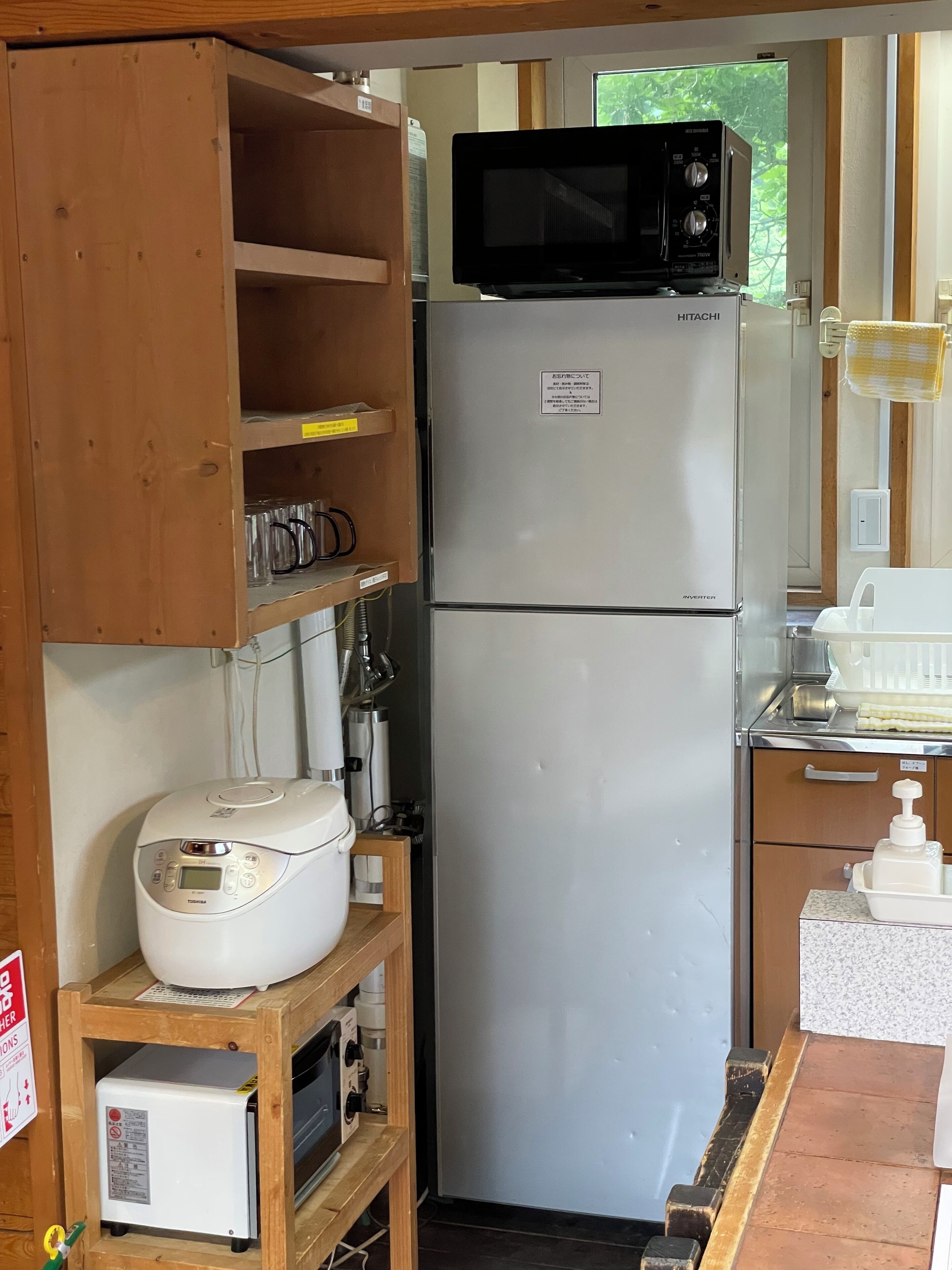 A compact kitchen with a refrigerator, microwave, and a rice cooker on a wooden shelf.