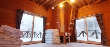Ein Zimmer in einer Blockhütte mit Holzwänden und schrägverdachter Decke. Zwei große Fenster mit Vorhängen, ein Stapel Decken und eine an der Wand angelehnte Leiter sind zu sehen.