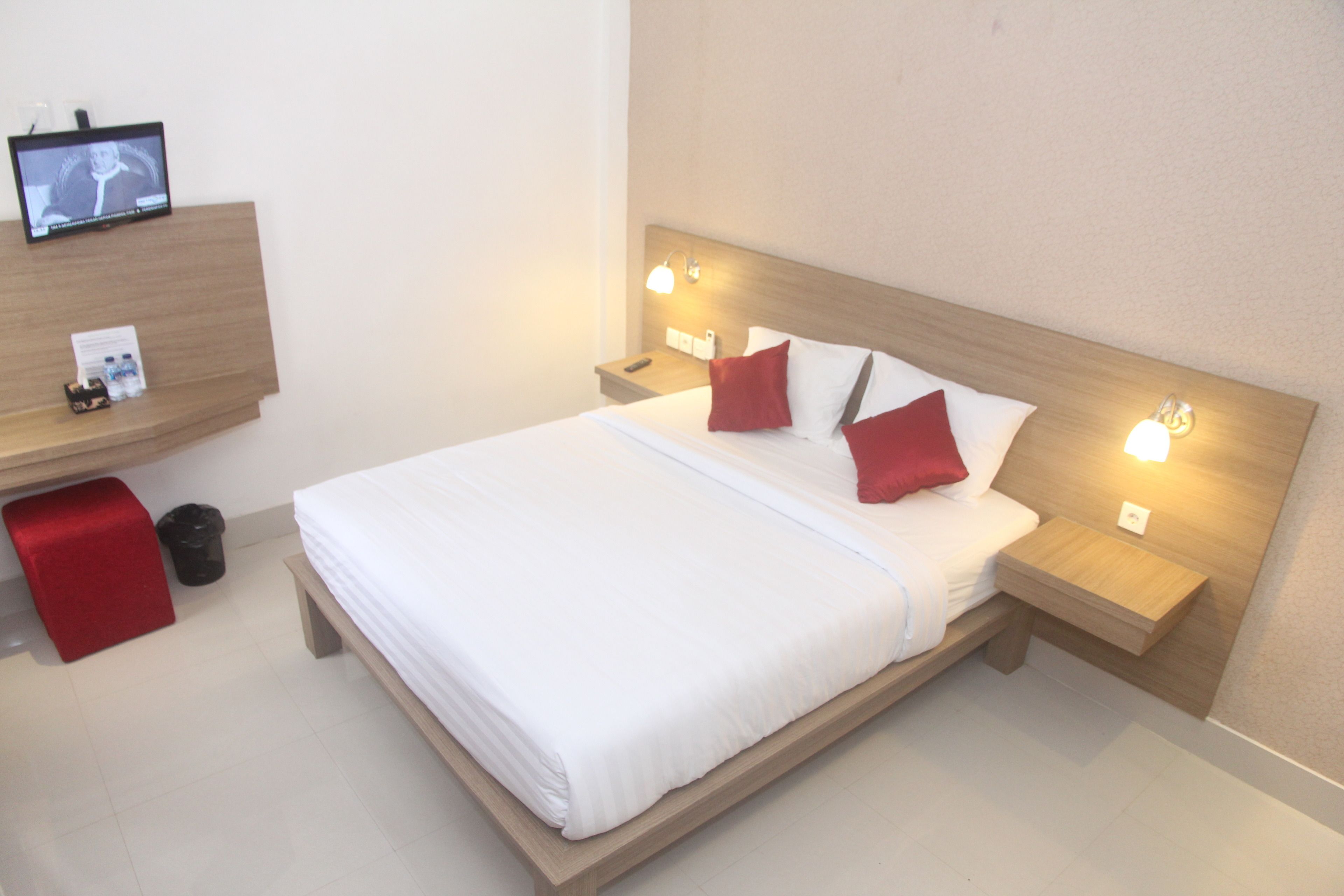 Una habitación de hotel con una cama, dos mesitas de noche, un televisor en la pared y un reposapiés rojo.