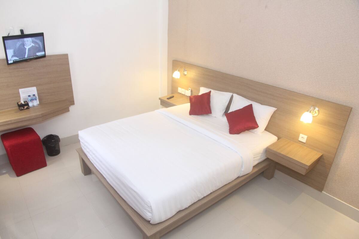 Une chambre d’hôtel avec un lit, des oreillers rouges, un téléviseur posé sur une étagère et des lampes de chevet.
