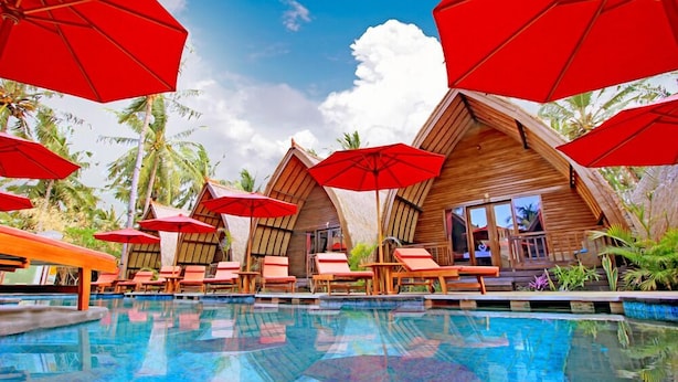 Un espace aménagé au bord de la piscine, avec des cabanons en bois, des parasols rouges et des chaises longues.