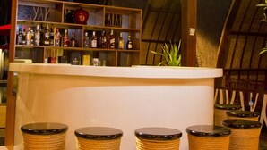 Un espace bar avec un plafond en bois et une structure de toit en chaume. On y trouve des tabourets en osier et un comptoir de bar avec des bouteilles.