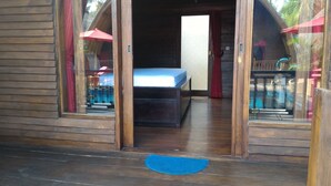 Une cabane en bois avec un lit, un tapis bleu et un parapluie rouge, visibles à travers les portes en verre.