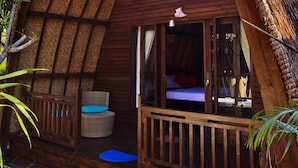 Un bungalow en bois avec un toit de chaume, un balcon agrémenté de meubles en osier et un petit espace extérieur aménagé pour s’asseoir.
