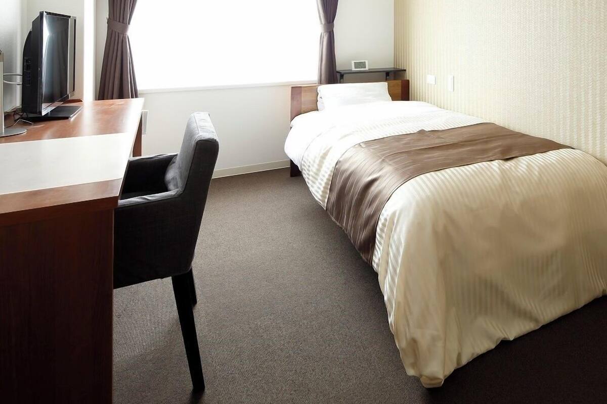 Quarto de hotel com cama, escrivaninha, cadeira, televisão e cortinas.