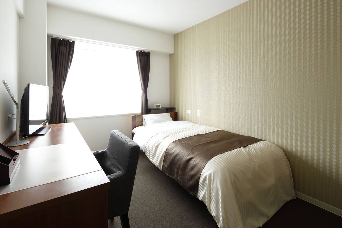 Quarto de hotel com uma cama, escrivaninha, cadeira, televisão e janela com cortinas.