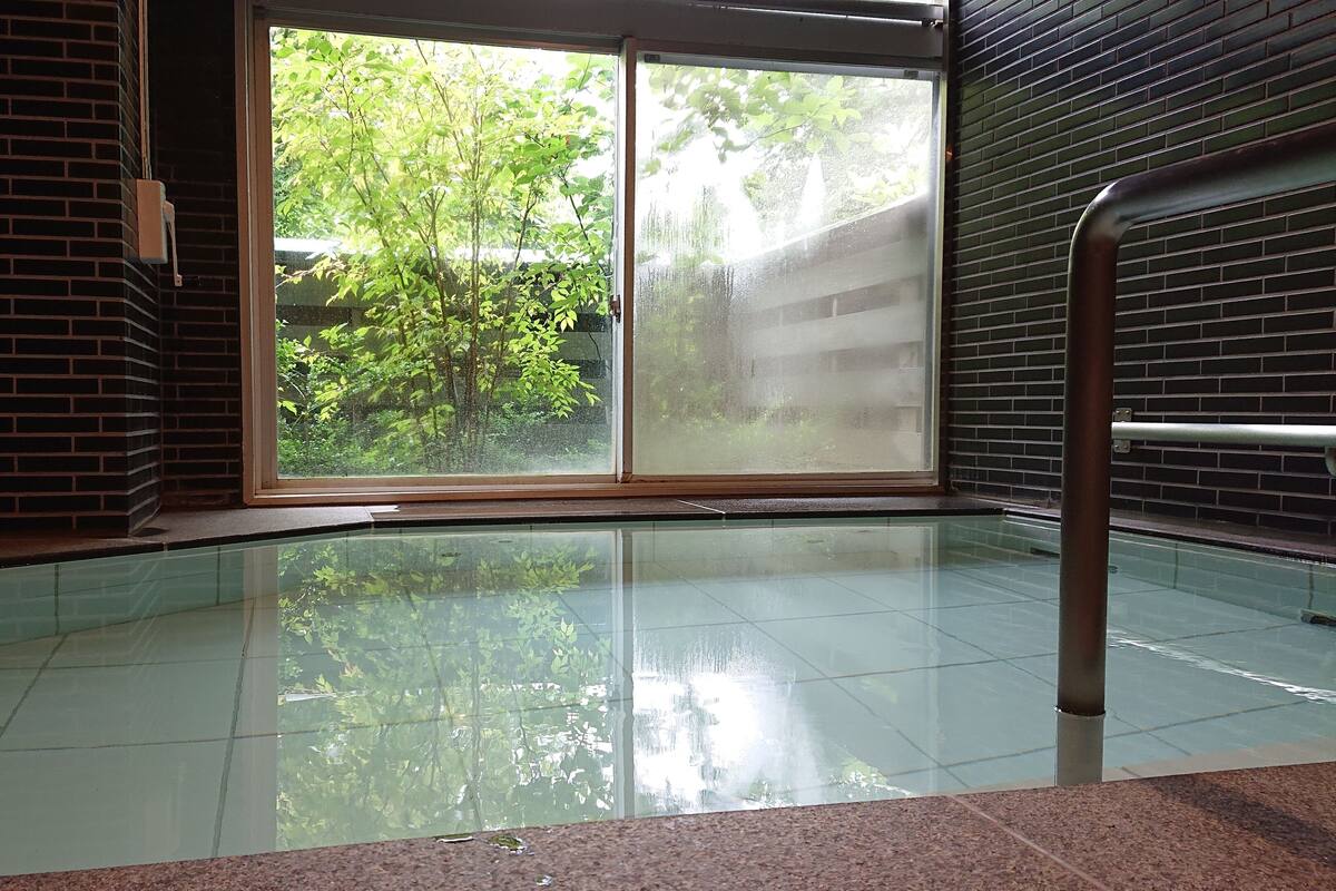 Piscina interior com piso de vidro, vista para o verde e parede de tijolos.