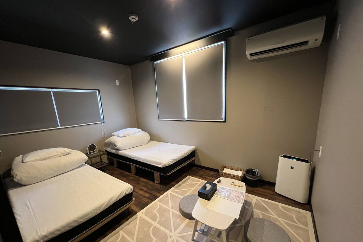 Quarto de hotel compacto com cama, mesa de cabeceira, ar condicionado e um pequeno espaço de estar com mesa e cadeiras.