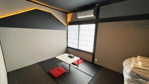 A compact room with a bed, a small table with a red cushion, a window with bars, and an air conditioning unit. - Hostel&Information SAMURISE (Fujikawaguchiko)
