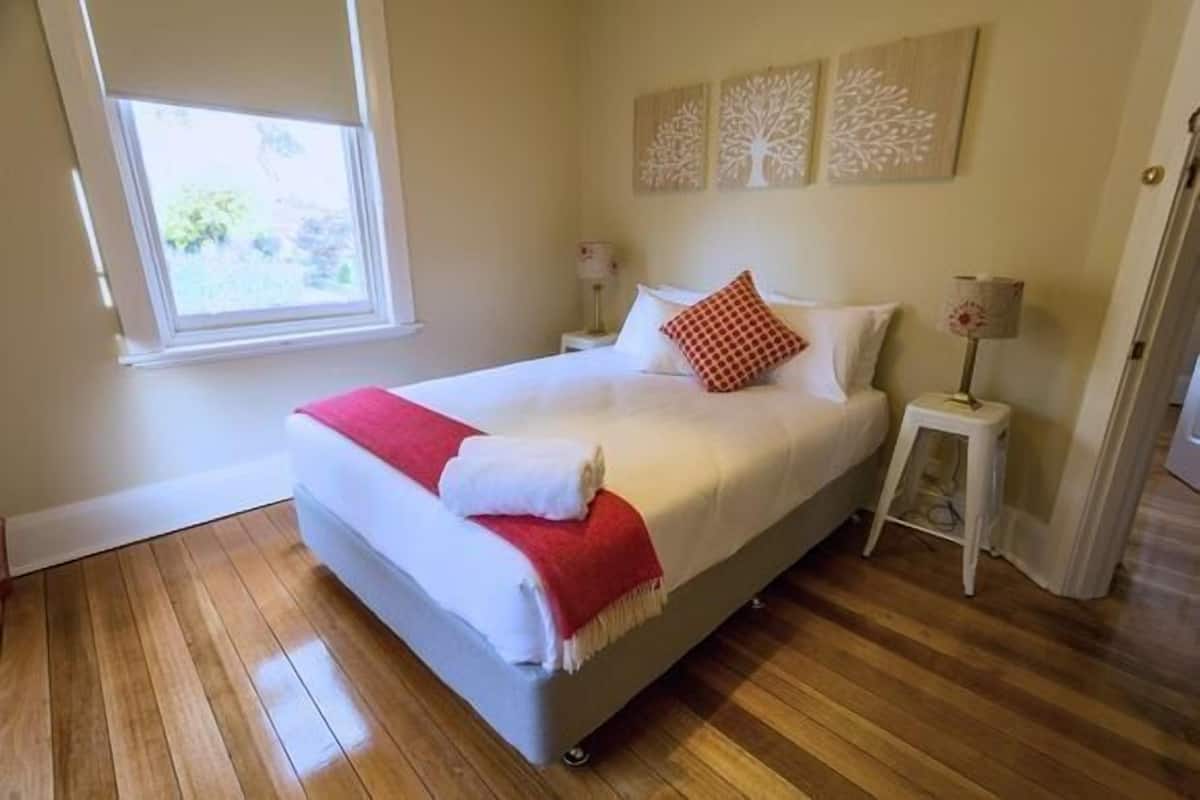 A neatly made bed with a red throw blanket, wooden flooring, and a window with a view of greenery.