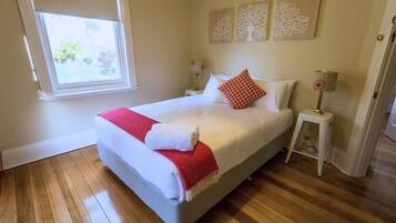 A neatly made bed with a red throw blanket, wooden flooring, and a window with a view of greenery.