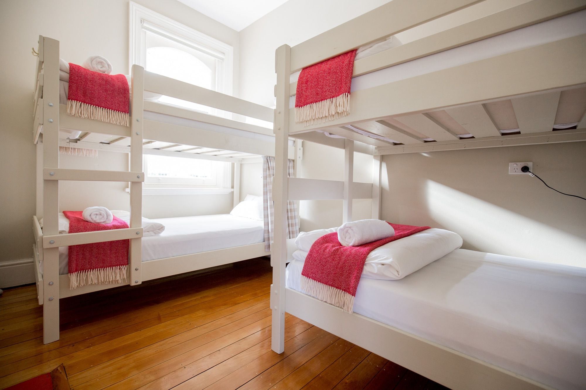 Dos literas con ropa de cama blanca y mantas rojas, suelo de madera y una ventana que deja entrar luz natural.