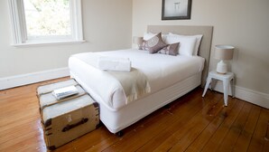 Una cama bien hecha con una colcha blanca y almohadones, una lámpara de noche, una maleta y un cuadro en la pared.