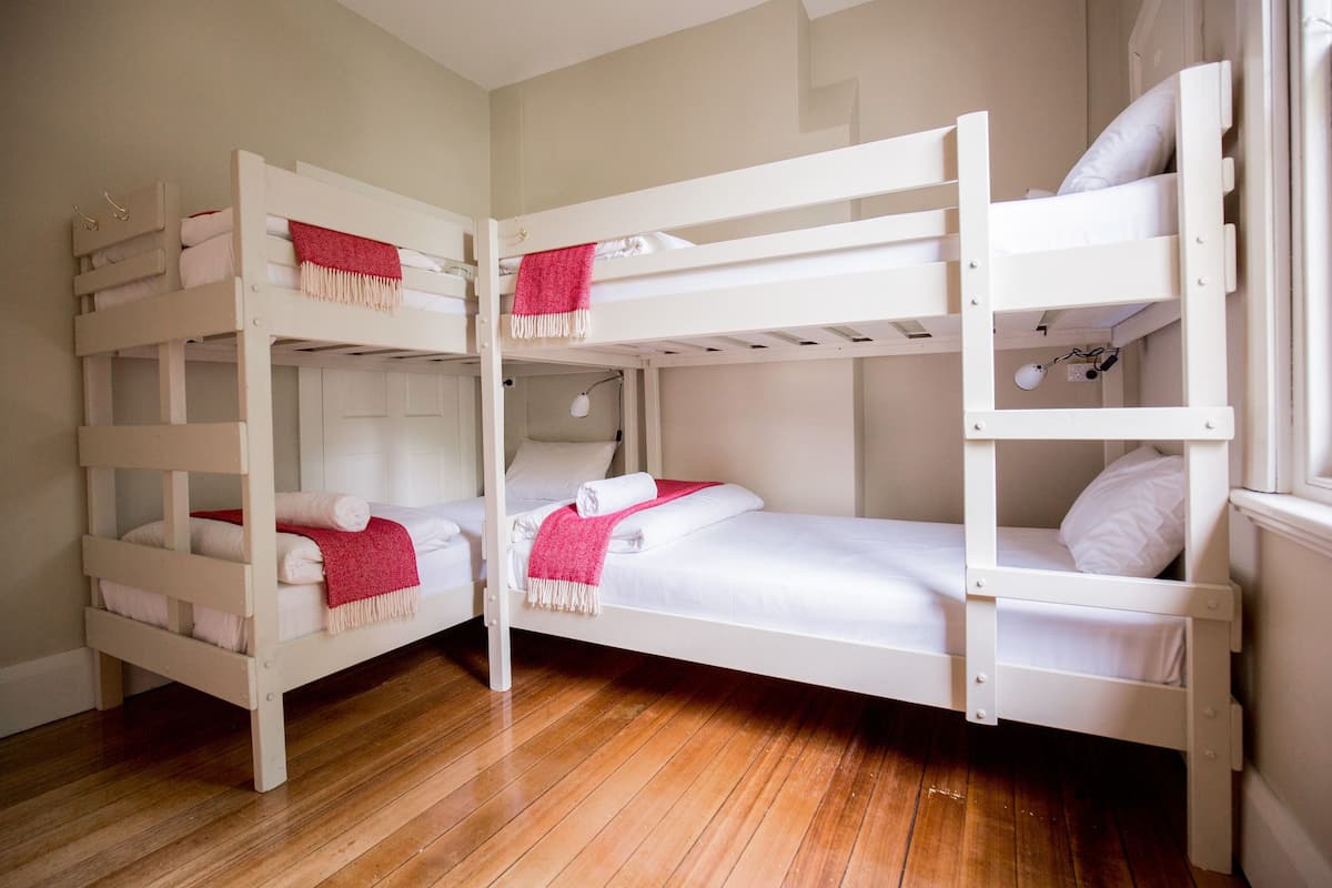 A bunk bed room with white beds and red blankets, wooden flooring, and a window on the right.