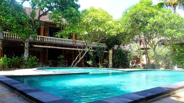 A swimming pool surrounded by lush greenery and traditional architecture. - Budhi Beach Inn (Kuta)