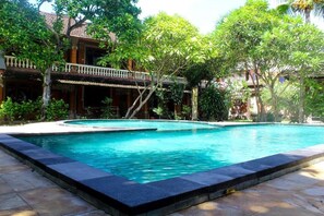 A swimming pool surrounded by lush greenery and traditional architecture. - Budhi Beach Inn (Kuta)