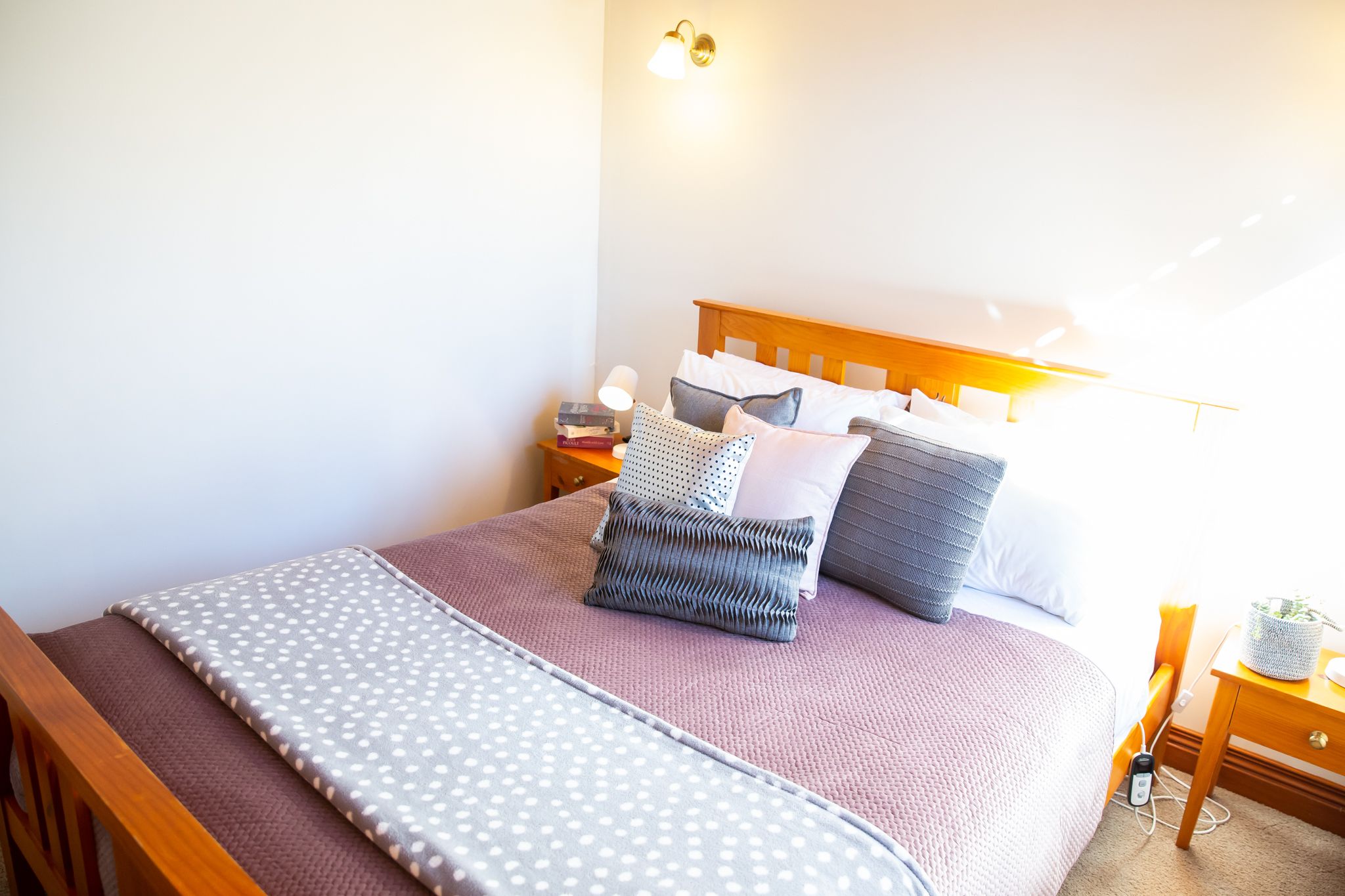 Una cama bien hecha con cubrecama y almohadas estampadas, una mesita de noche con lámpara y un mueble auxiliar con libros.